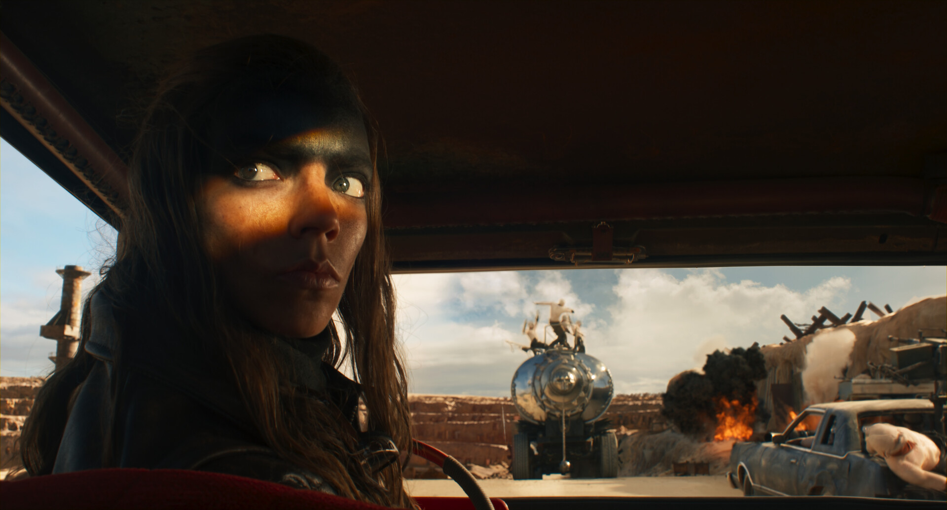Anya Taylor-Joy gazes intensely from the driver’s seat in a desert action scene. In the background, a large vehicle explodes, with another car and a truck-mounted machine gun visible amid the chaos.