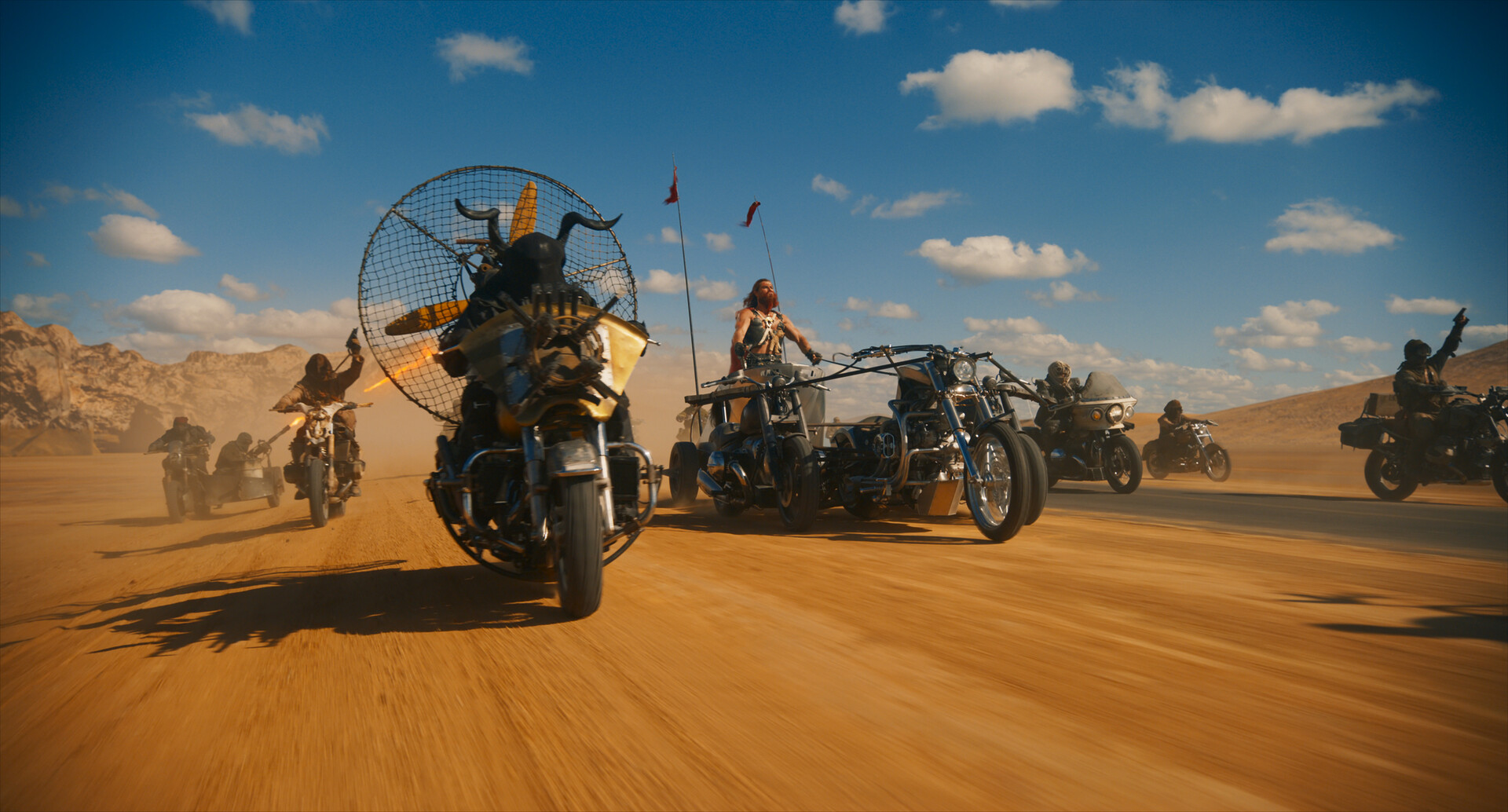 Chris Hemsworth leads a group of riders on customized motorcycles and trikes across a dusty desert. Each vehicle is uniquely outfitted with makeshift armor and decorations, emphasizing a rugged, post-apocalyptic style. The riders speed across the sandy terrain under a clear blue sky, stirring up a trail of dust.