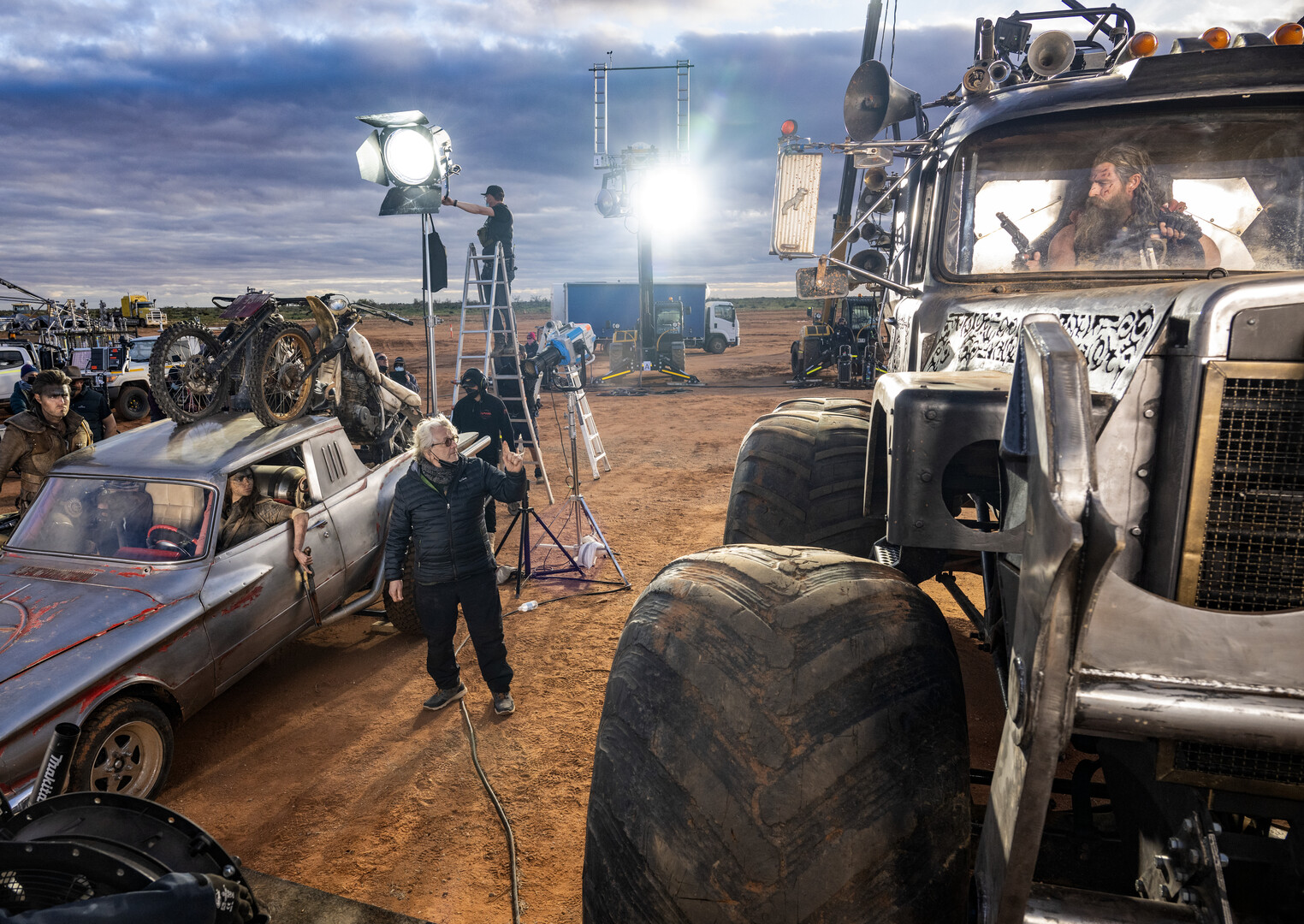 Behind-the-scenes on a movie set showcasing a bustling atmosphere with key cast and crew. Featured are actors Tom Burke and Anya Taylor-Joy in a car, director George Miller instructing near the camera, and Chris Hemsworth visible through a truck window. The scene is filled with film equipment like cameras and lights, set against a backdrop of vehicles and a cloudy sky, highlighting the extensive production setup.