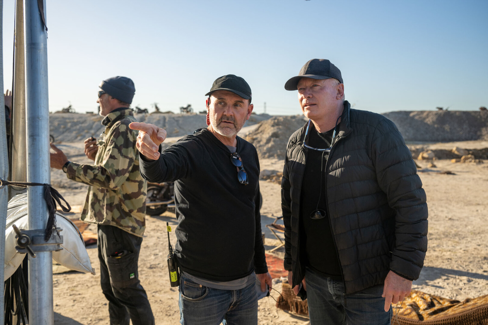 Two men discussing on a film set, with one pointing and explaining something in the distance. They are dressed casually, one in a black long-sleeve shirt and the other in a padded black jacket. In the background, film crew members work around bikes and equipment in a sandy, open outdoor location under a clear sky.
