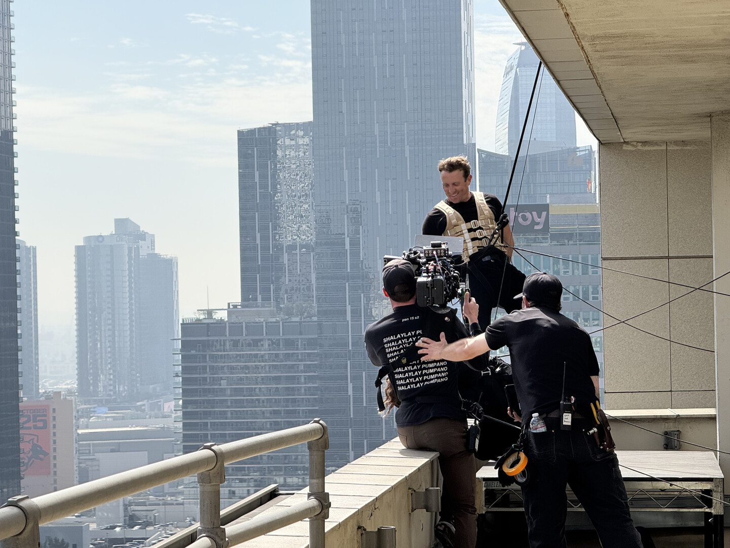 A stunt performer wearing safety gear and film crew on the balcony of a skyscraper